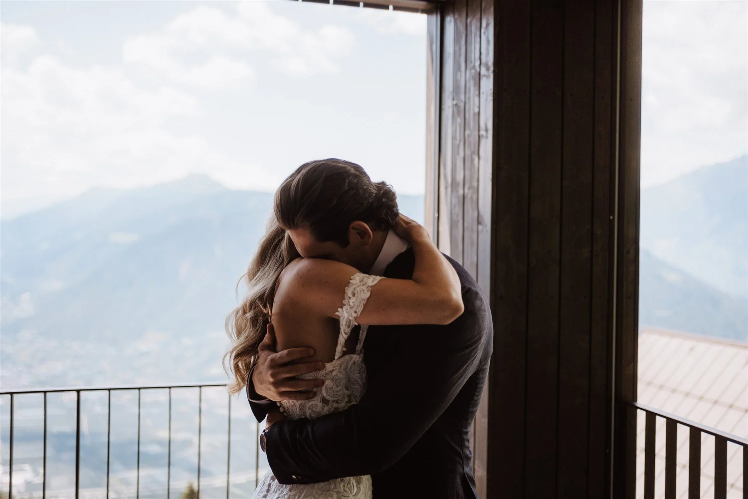 A couple sharing an embrace, with the woman in a white laced dress and the man in a dark suit, standing on a balcony overlooking mountains and sky, during daytime.