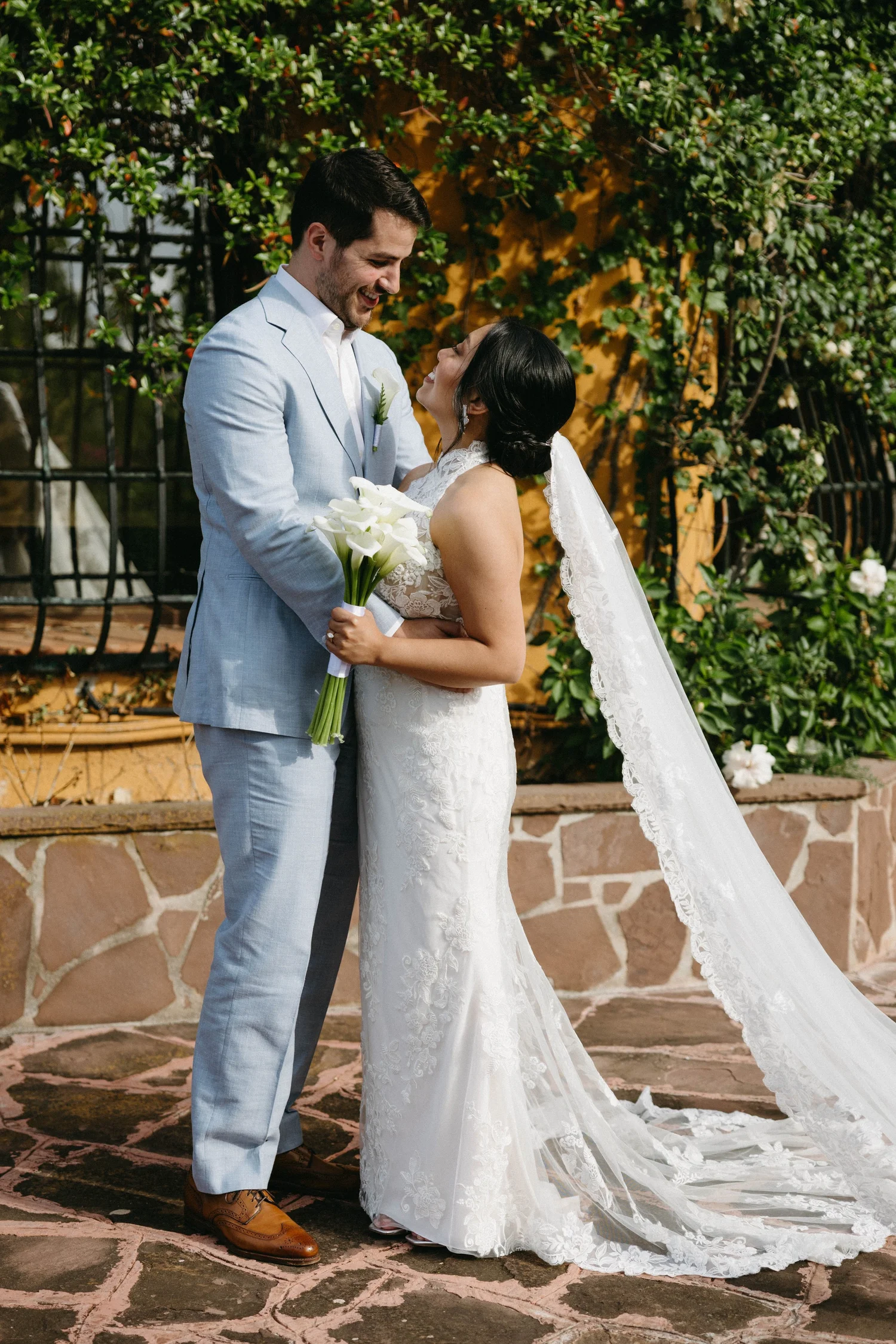 A bride and groom holding hands and smiling at each other outside, with the bride holding a bouquet of white calla lilies, surrounded by green foliage and a decorative stone wall.