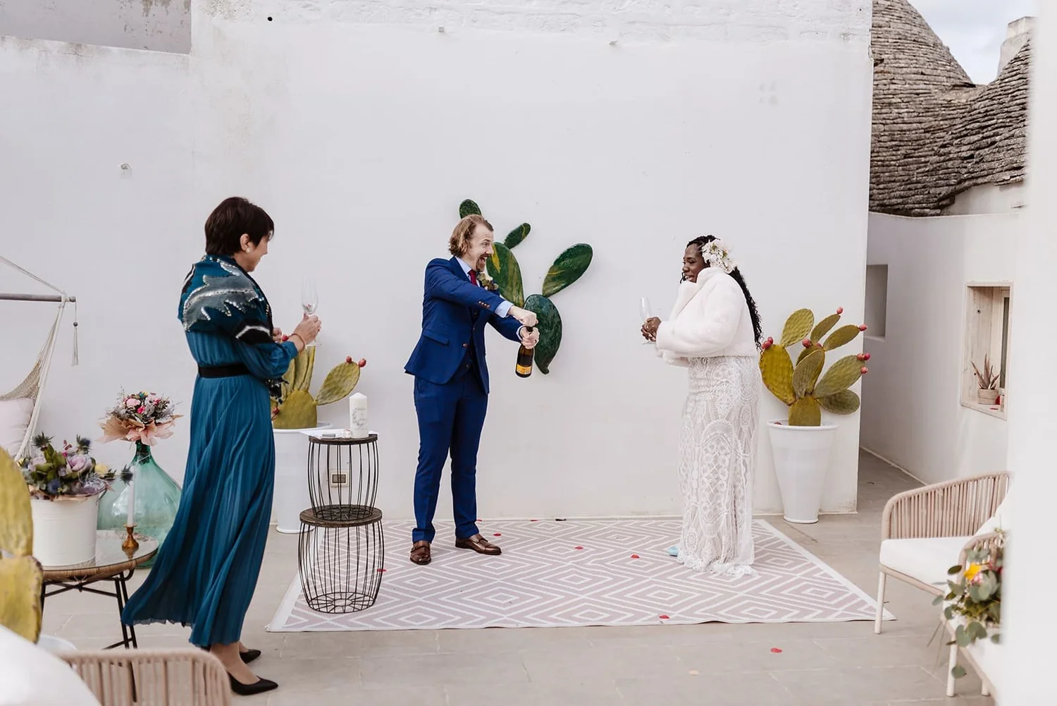 A wedding ceremony with three women, one of whom is a bride, dressed in a lace wedding gown, holding a glass, and adorned with flowers in her hair. The man in a blue suit and tie is opening a bottle of champagne. One guest is dressed in a long blue d
