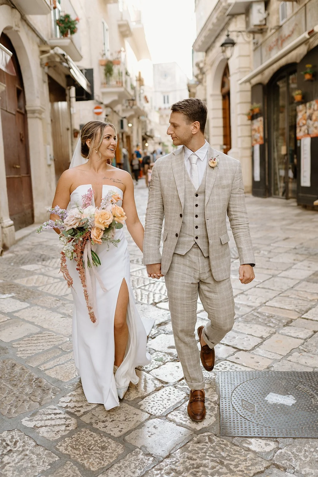 A bride and groom holding hands and walking on a stone-paved street in a European town, dressed in wedding attire. The bride wears a white gown with a thigh-high slit and holds a colorful bouquet, while the groom wears a light plaid suit with brown s