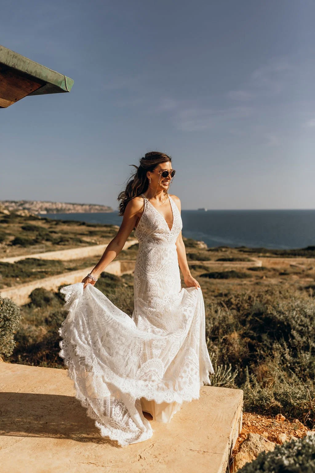 A woman in a white lace dress standing outdoors near the coastline under a clear blue sky, holding her dress with both hands and smiling.