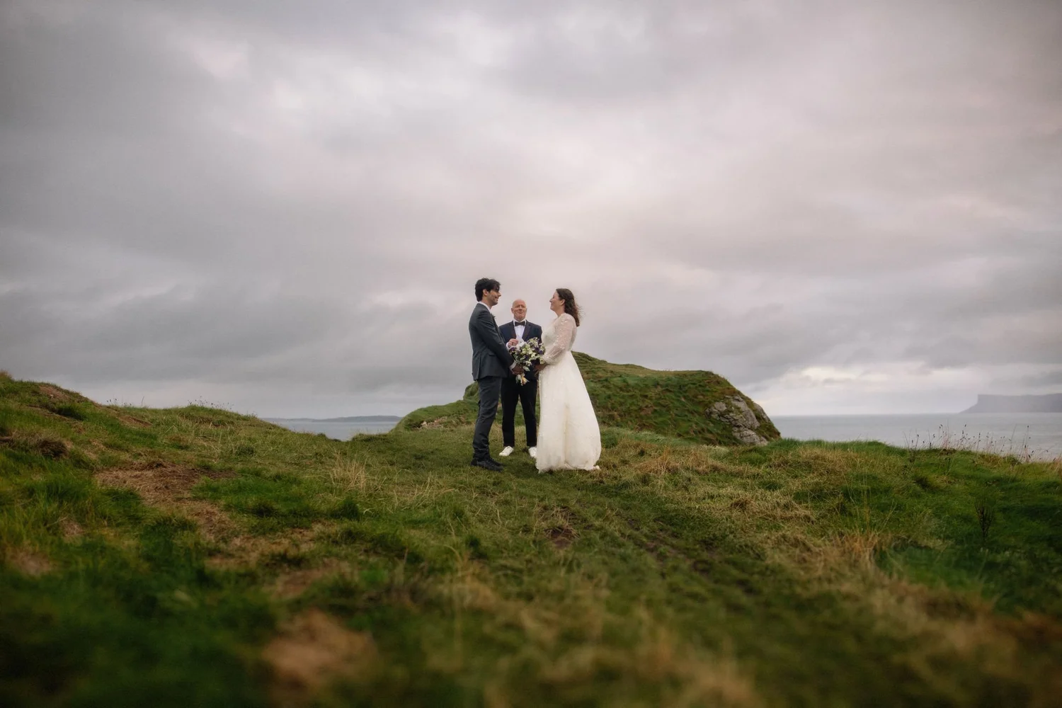 A bride and groom stand holding hands, facing each other, with an officiant in the middle outdoors on a grassy hill under a cloudy sky.