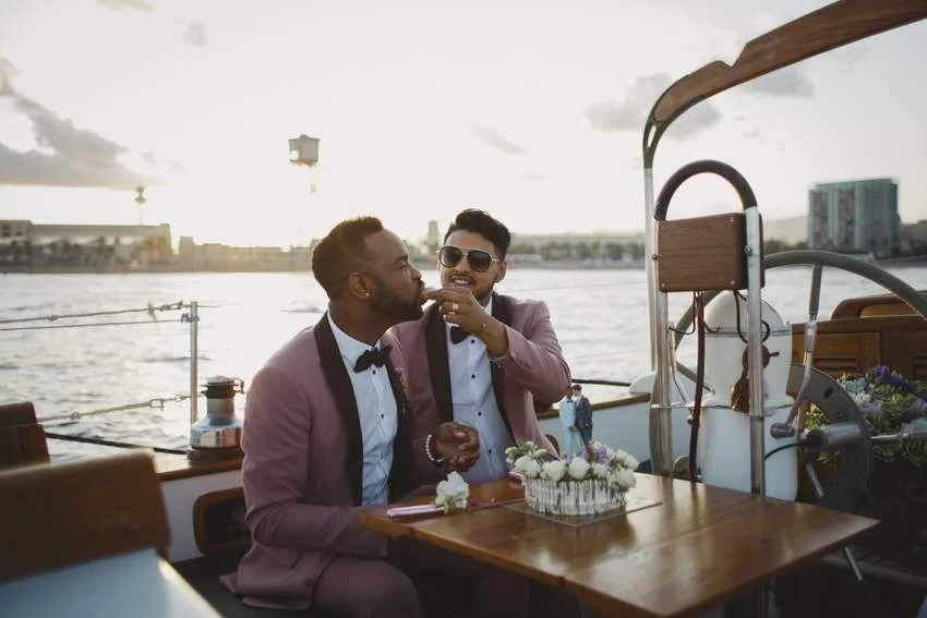 Two men dressed in suits with bow ties are sitting on a boat with a city skyline in the background. One man is feeding cake to the other, both smiling and enjoying the moment during sunset.