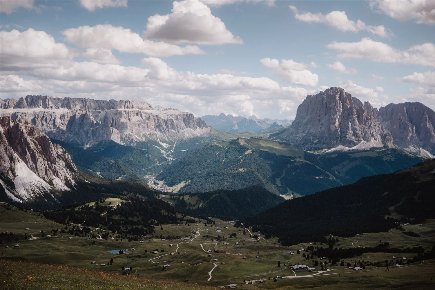 A scenic mountain landscape with tall, rugged gray peaks under a partly cloudy sky. Below the mountains, there are green valleys with scattered small houses, winding roads, and patches of forest.