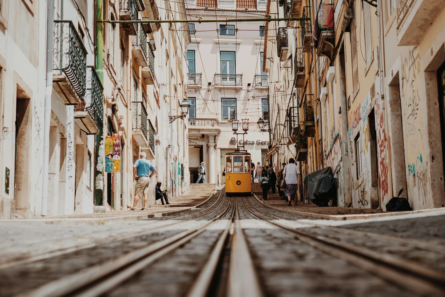 View of a Lisbon street with funicular train, people walking, colorful buildings with balconies, and overhead wires for the tram.