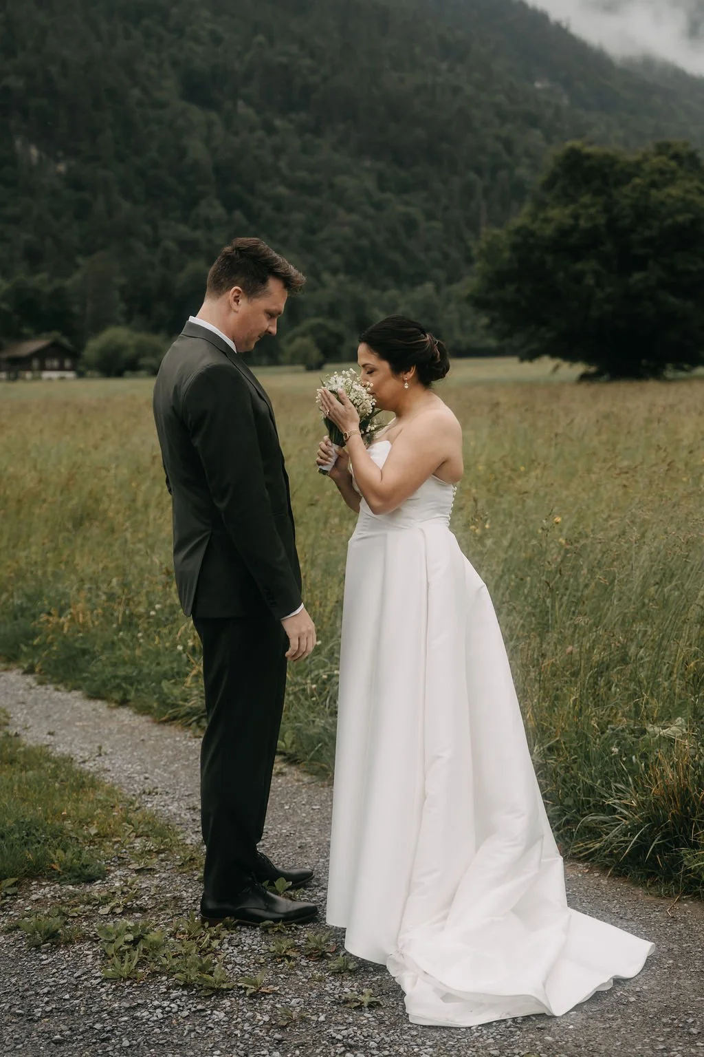 A bride and groom standing on a dirt path in a field with mountains in the background, the bride holding a bouquet and smiling while the groom looks down.