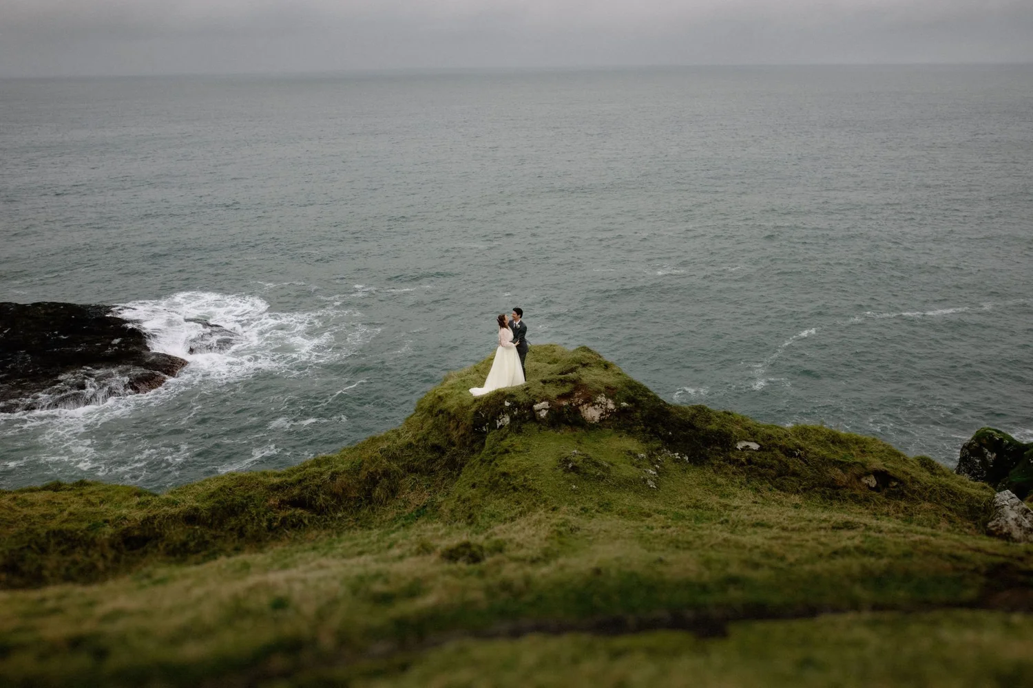 A bride and groom standing on a grassy cliff edge overlooking the ocean, holding hands.