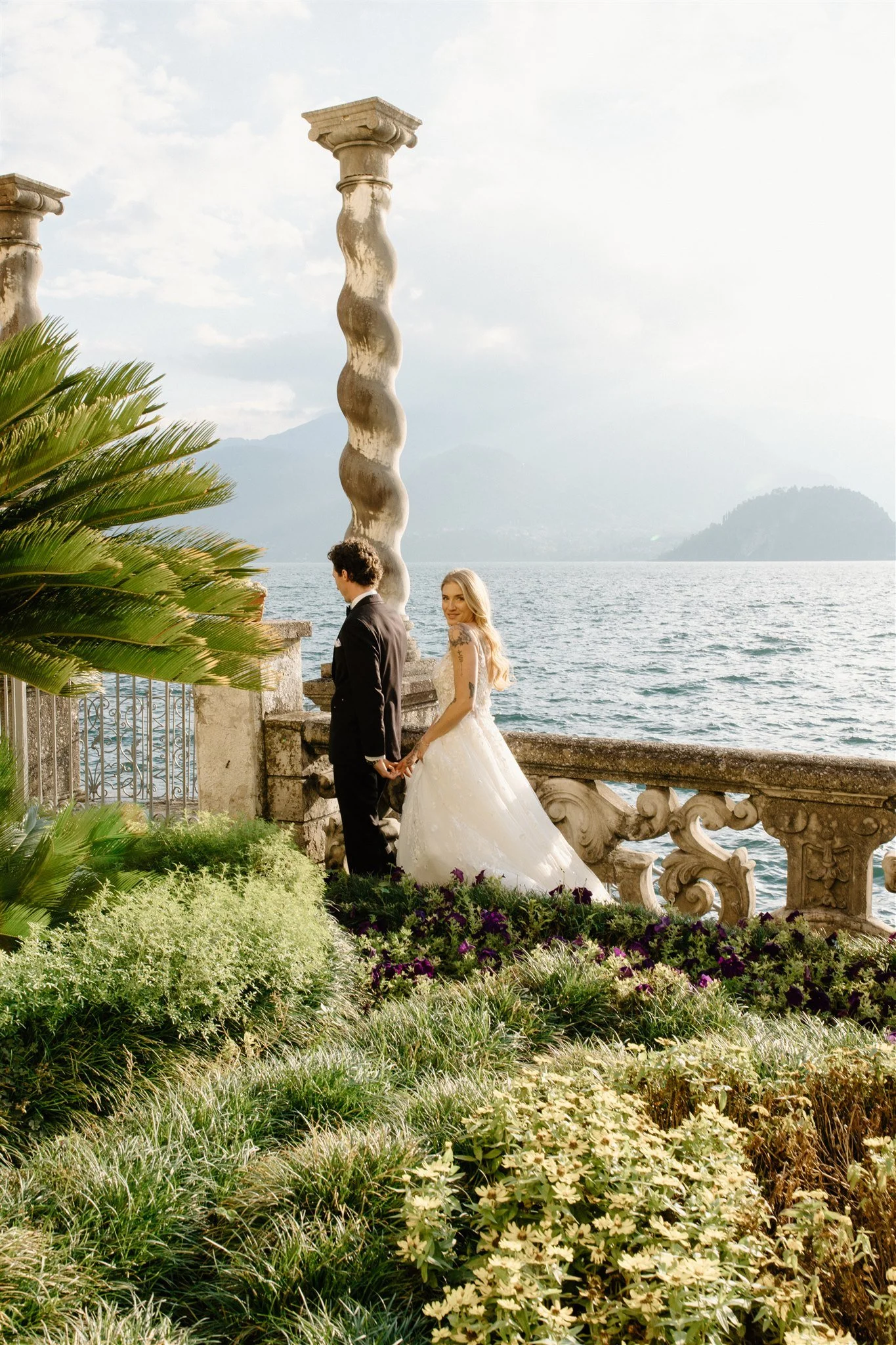 A bride and groom holding hands by a lake with scenic mountains in the background, surrounded by flowers and greenery.