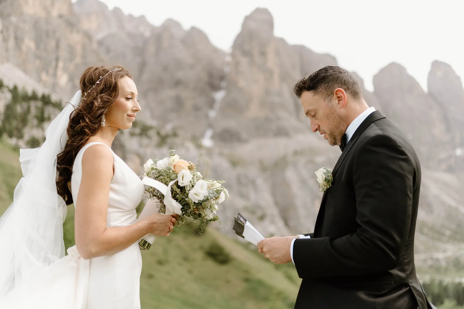 A bride and groom exchanging vows outdoors, with the bride holding a bouquet and the groom reading from a paper, mountains in the background.