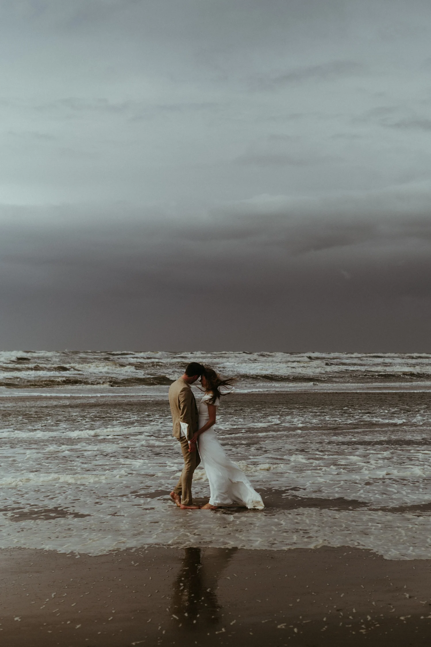 A couple dressed in wedding attire standing on the beach, kissing with ocean waves in the background and dark, cloudy sky above.