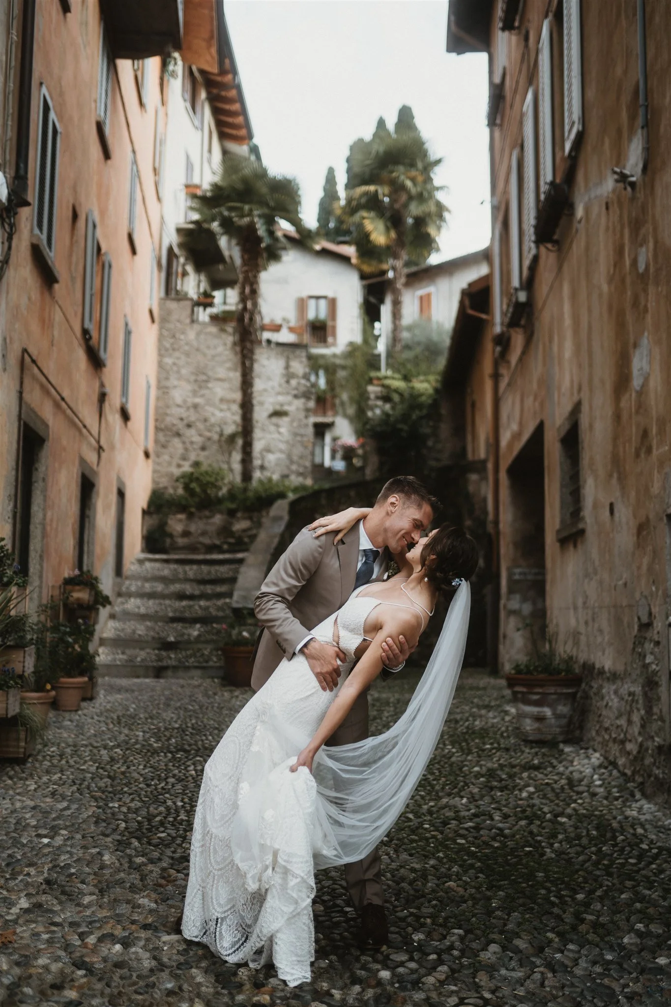 A bride and groom sharing a romantic dance in an outdoor, cobblestone alleyway surrounded by rustic buildings and palm trees.