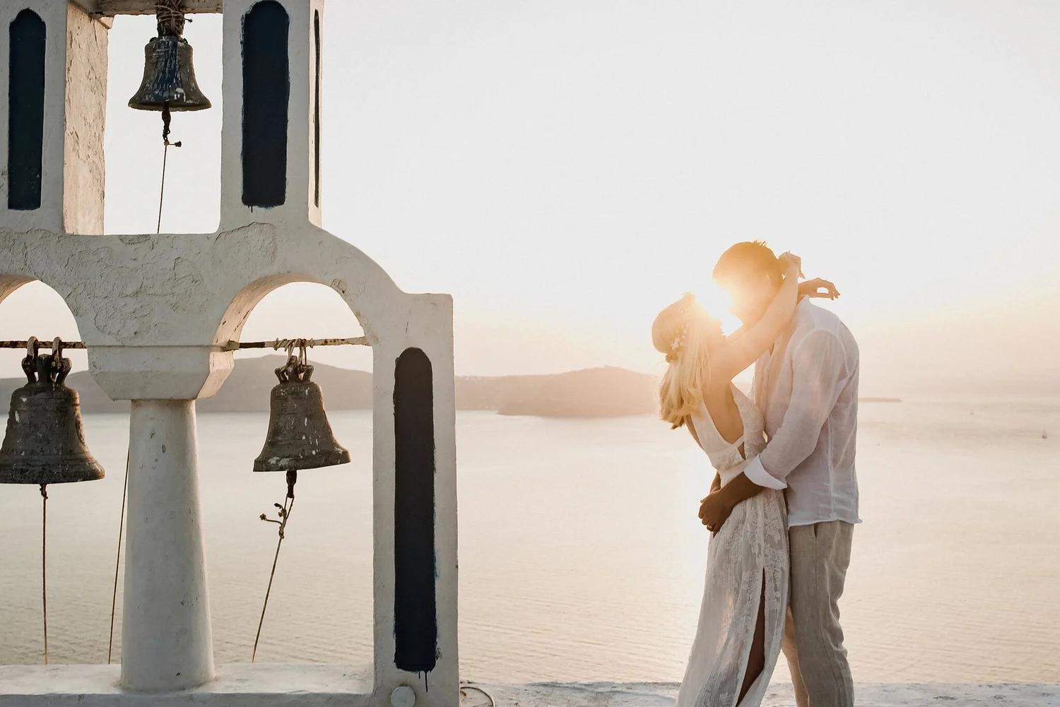 A couple stands close together, embracing each other with their faces near, backlit by the setting sun on a beach. They are dressed in light, flowy clothing, with the woman wearing a white dress and the man in light-colored trousers and a shirt. Behi