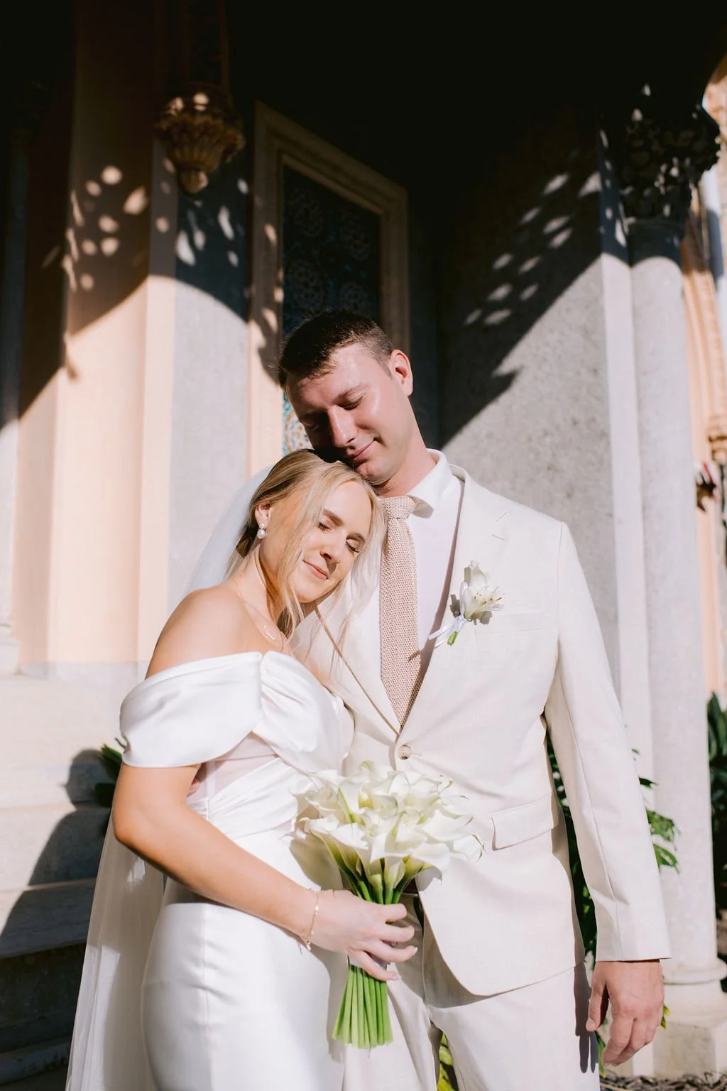A bride and groom embrace outside a building, with the sun casting shadows. The bride has blonde hair and is holding a bouquet of calla lilies. The groom has short dark hair and is wearing a light-colored suit with a tie and boutonniere.
