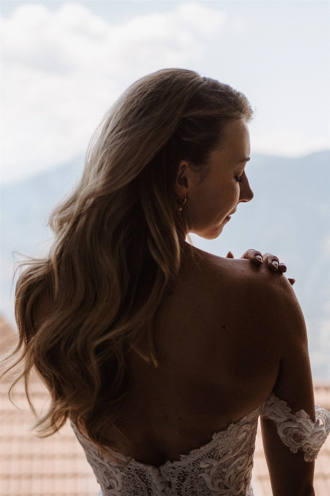 A woman with long, wavy blonde hair wearing a white lace dress, standing outdoors with a cloudy sky and distant mountains in the background.