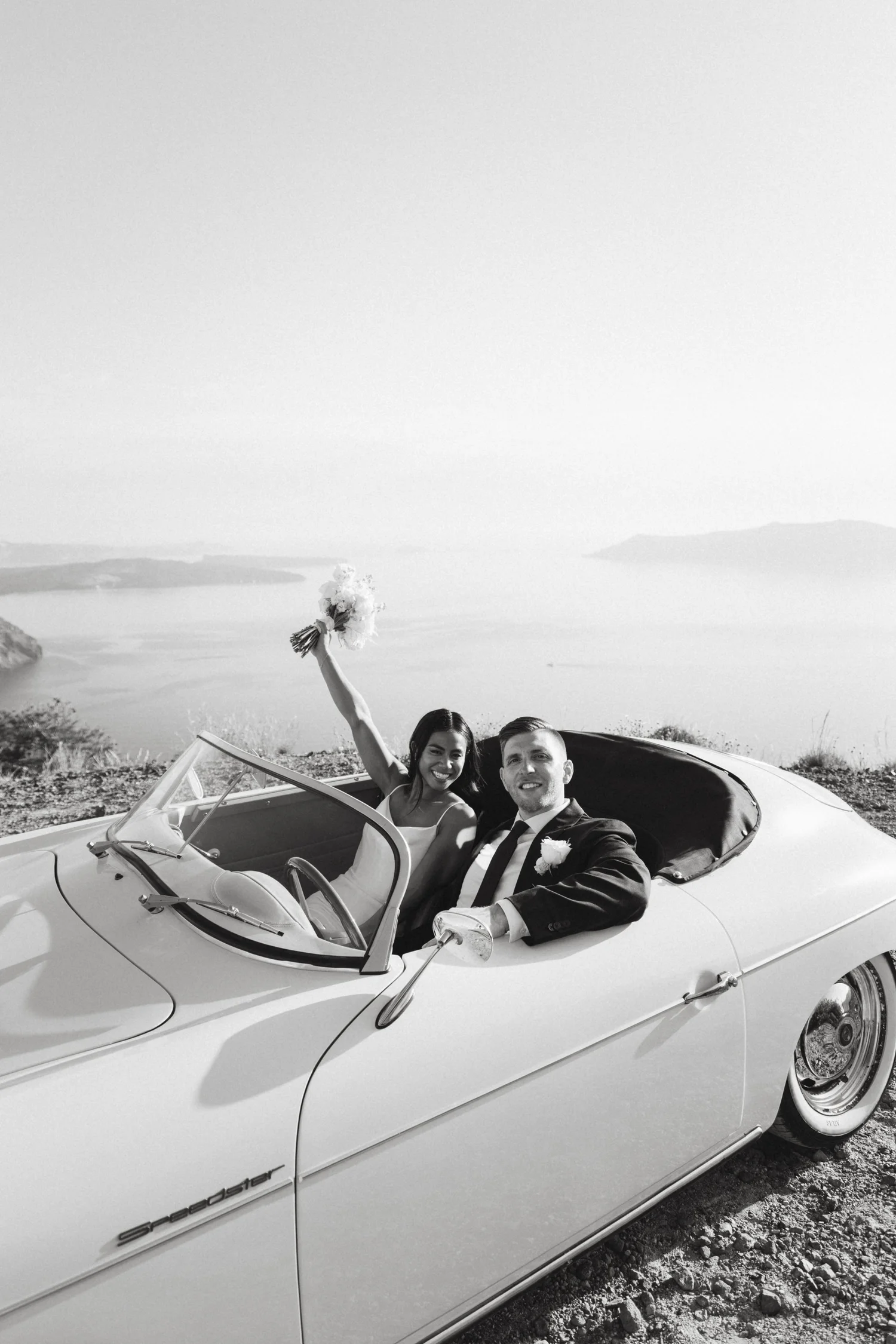Black and white photo of a man and woman in a vintage convertible car with hills and water in the background. The woman holds a bouquet of flowers and raises her arm in celebration. The man is dressed in a suit with a boutonniere, smiling at the came