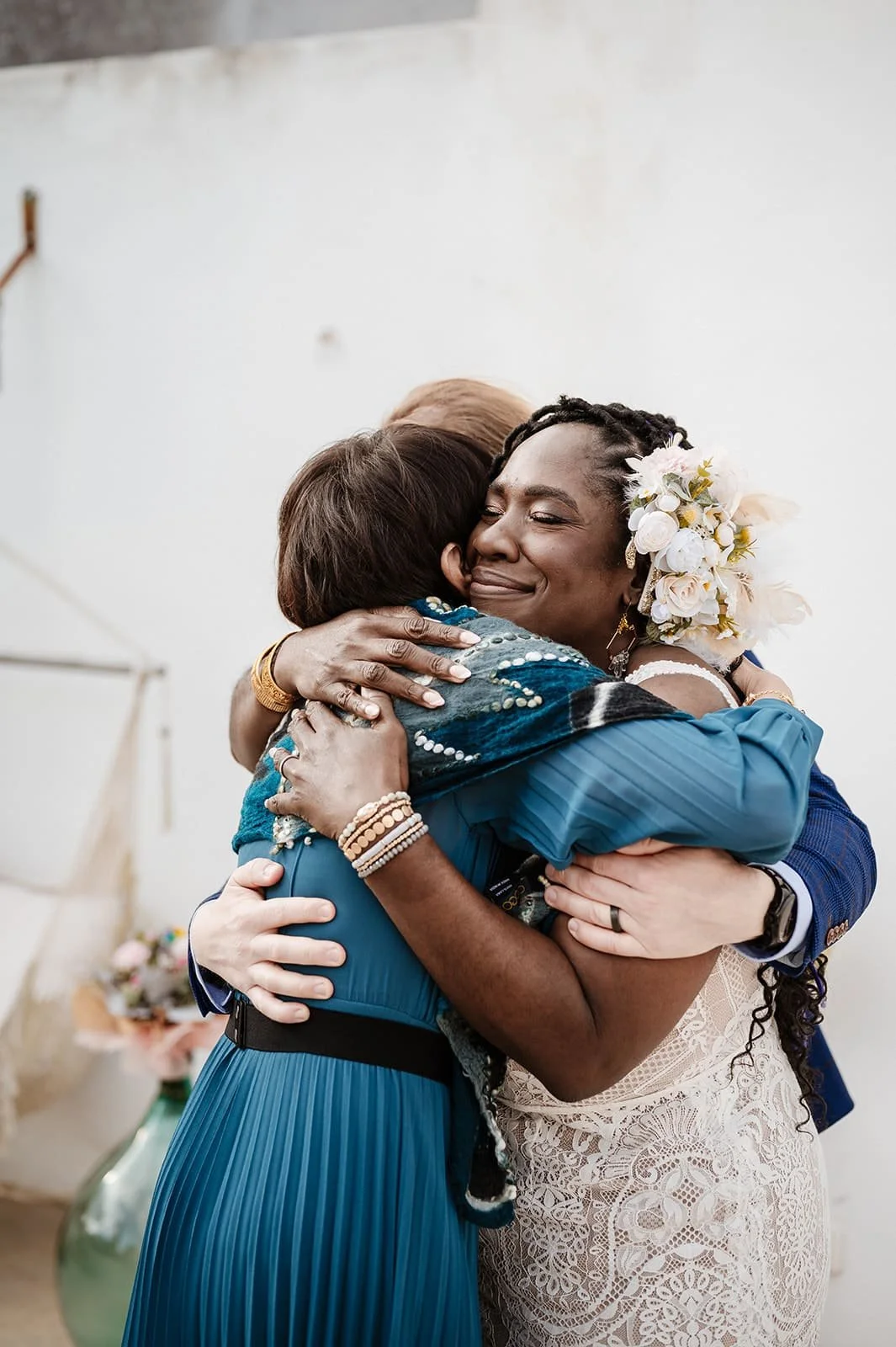 Two women hugging warmly at a celebration, one wearing a blue dress and the other in a white lace outfit with a floral headpiece.