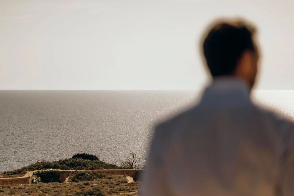 A blurry person standing outdoors in front of a large body of water and sky, with some vegetation in the foreground.