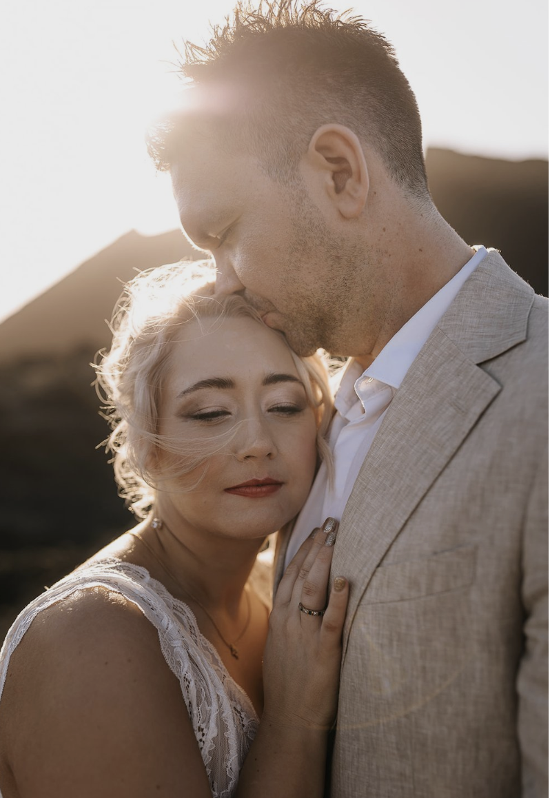A man and woman embrace in an outdoor setting during sunset, with the man kissing the woman's forehead and the woman with her eyes closed, wearing a white lace dress.