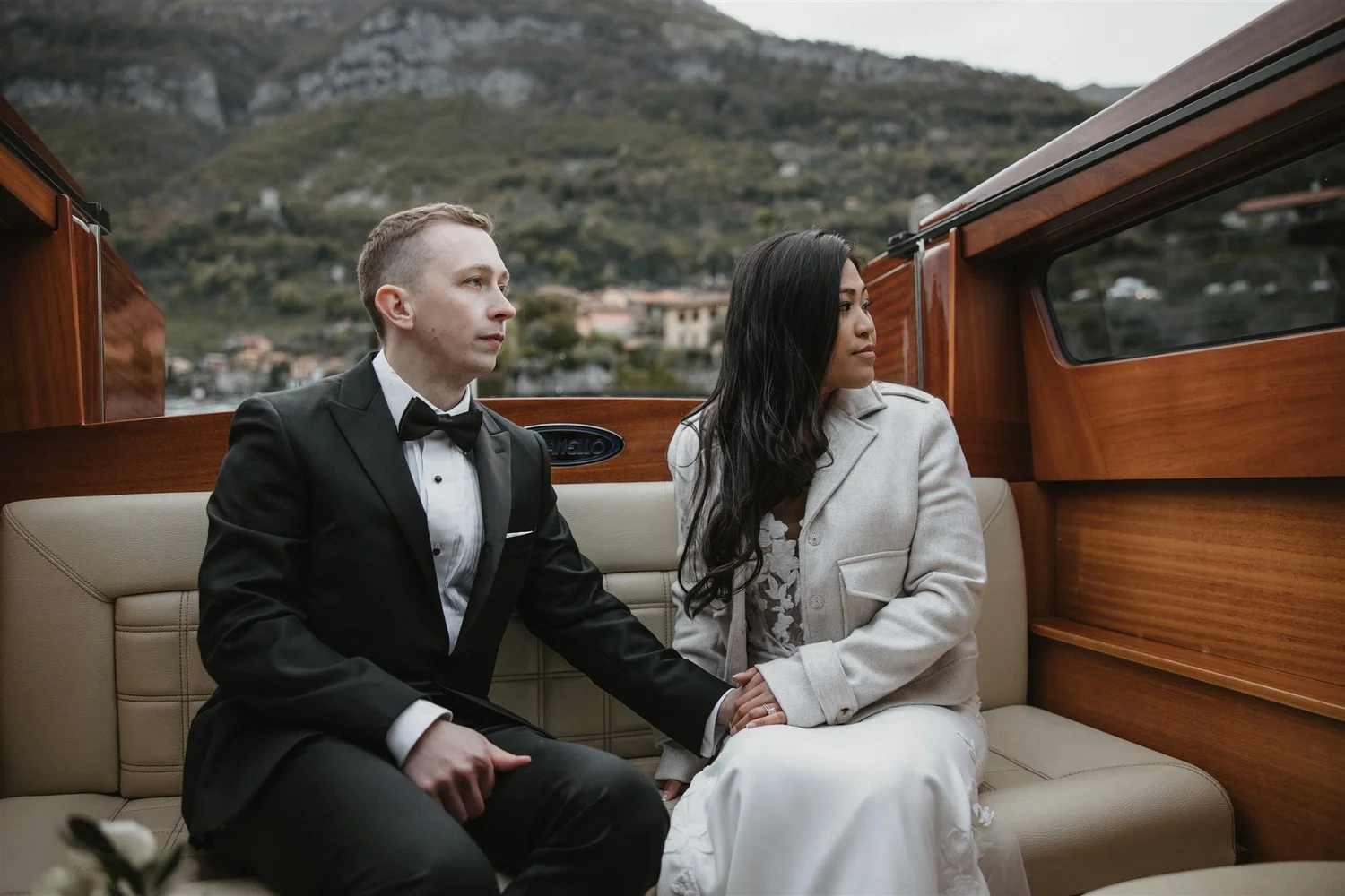 A man and woman sit closely together on a beige leather bench inside a boat, with their hands gently touching. The man is dressed in a black tuxedo with a bow tie, and the woman is wearing a white lace dress and a light-colored jacket. They look cont