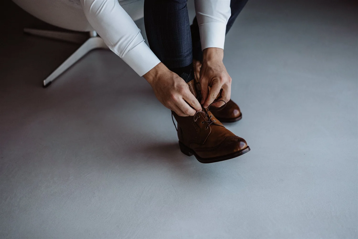 Person in white shirt and dark pants tying the shoelaces of brown leather boots while sitting on a white chair.