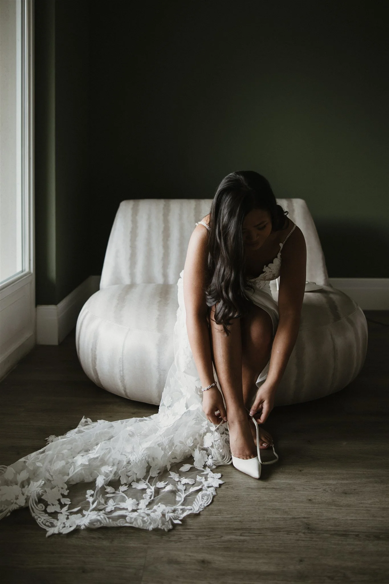 A woman in a wedding dress is sitting on a couch as she puts on white high-heeled shoes, with her wedding train and flower petals on the wooden floor in front of her. She is in a room with dark walls and a window to her left.