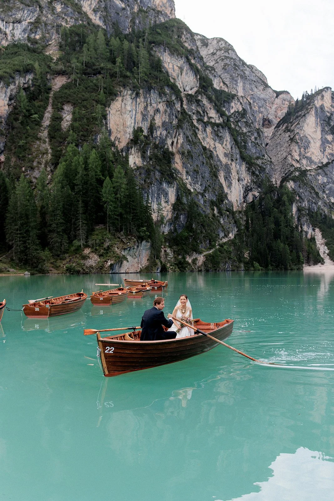 A couple in wedding attire in a rowboat on a turquoise lake, surrounded by tall green mountains and additional rowboats in the background.