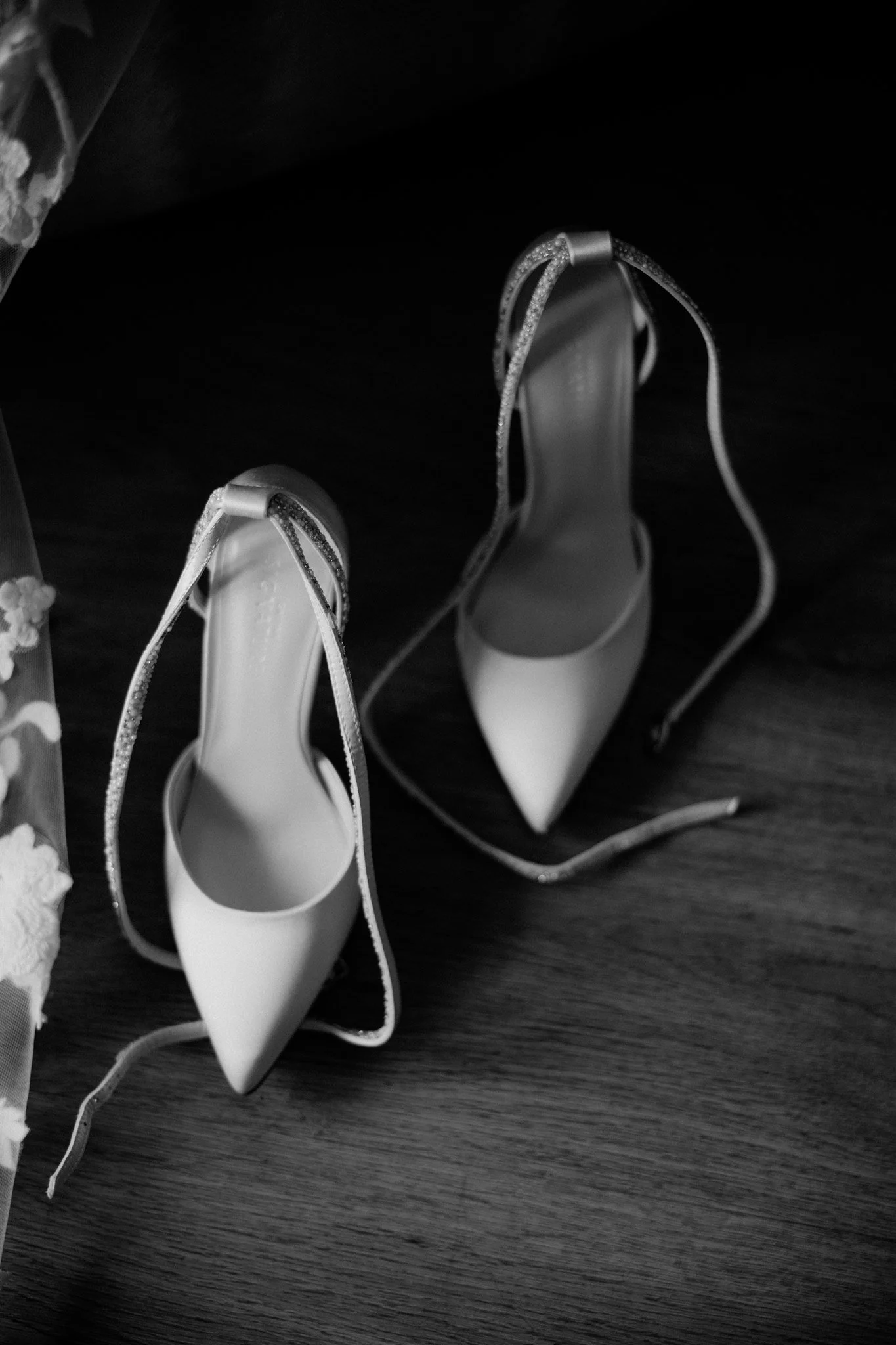 White high-heeled shoes with ankle straps and rhinestone embellishments on a dark wooden surface.