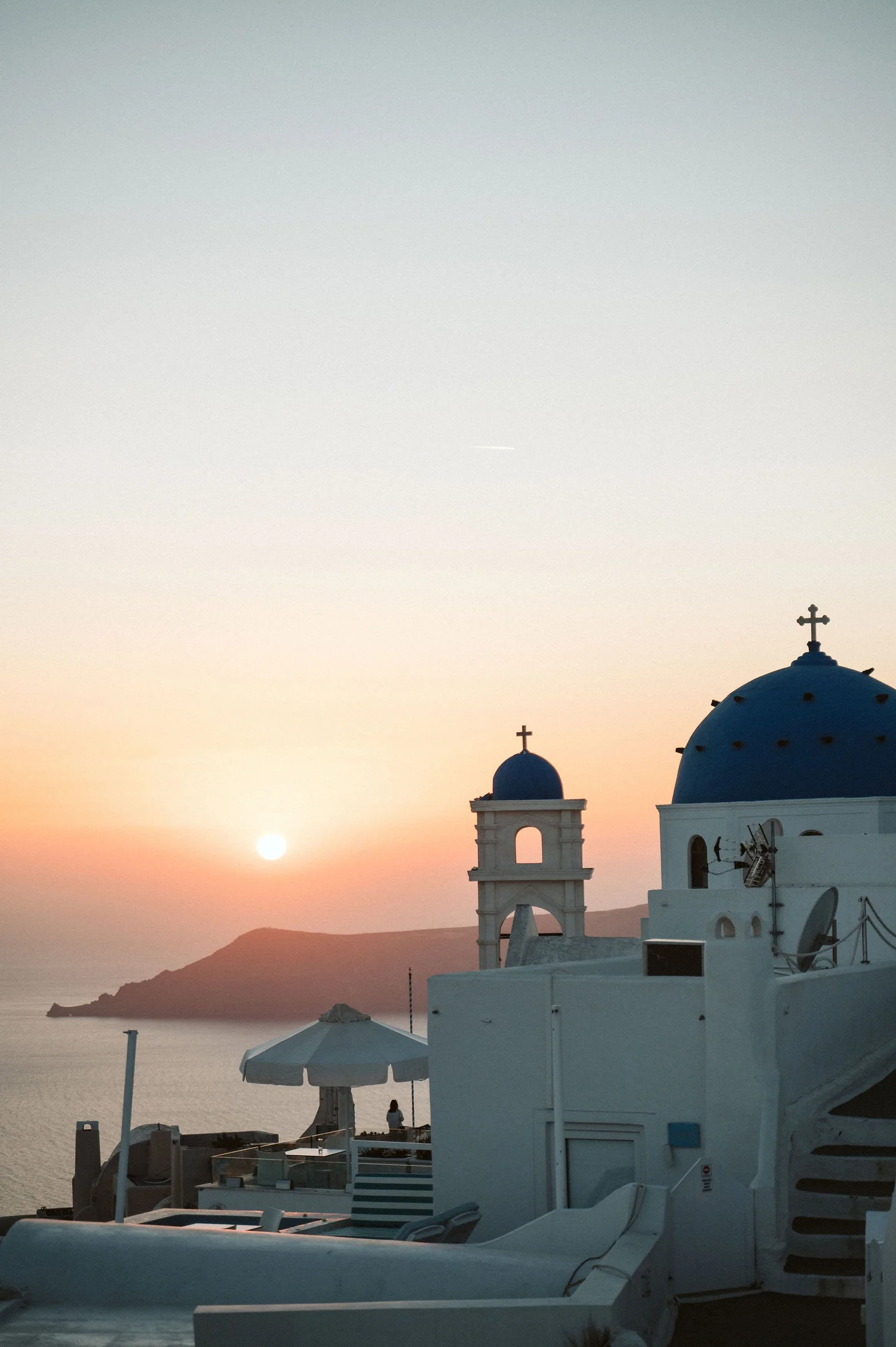 Sunset over a white-washed Greek church with blue domes and crosses, overlooking the sea, with a person sitting under an umbrella on the terrace.