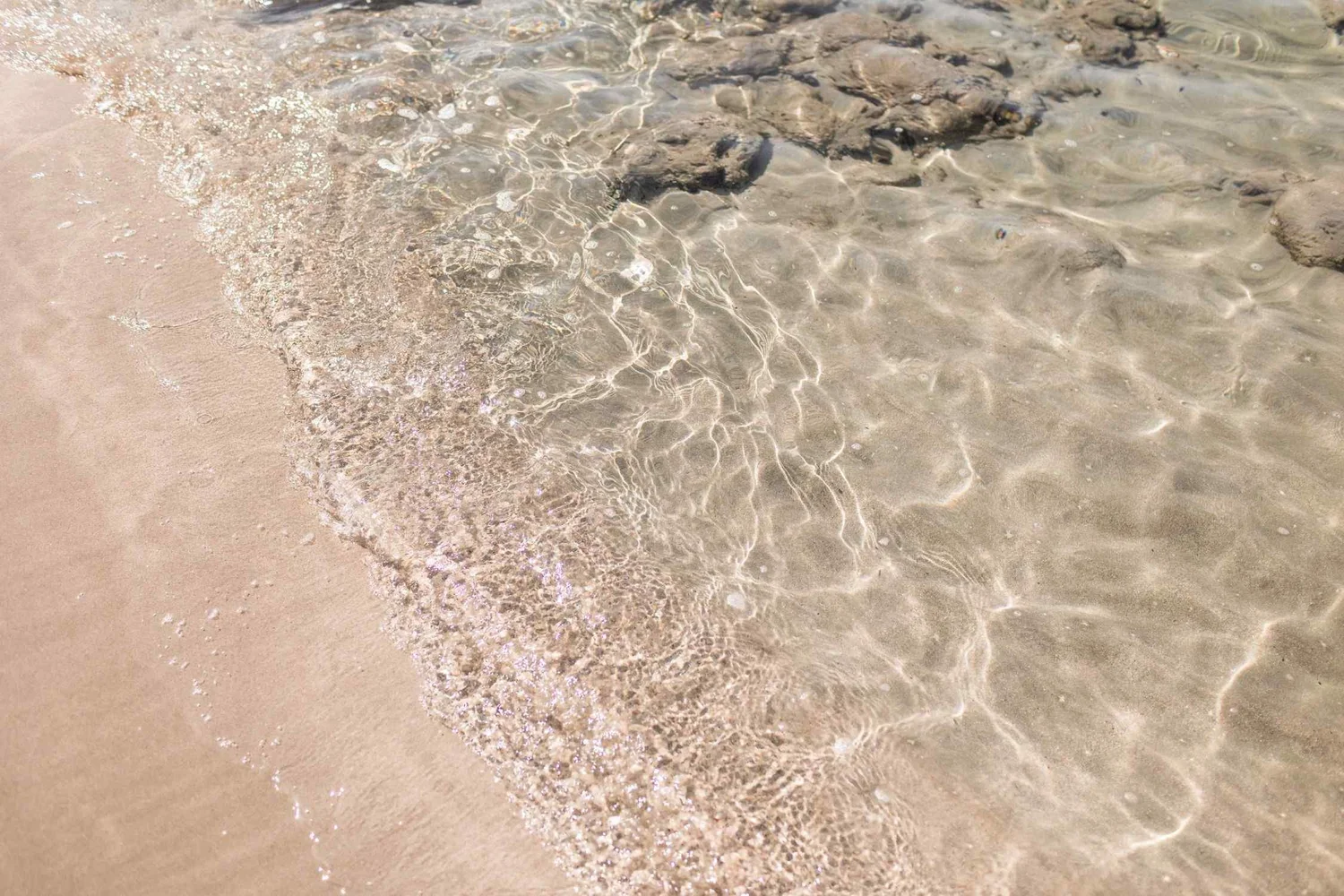 Clear shallow water lapping onto a sandy beach with some rocks visible beneath the surface.