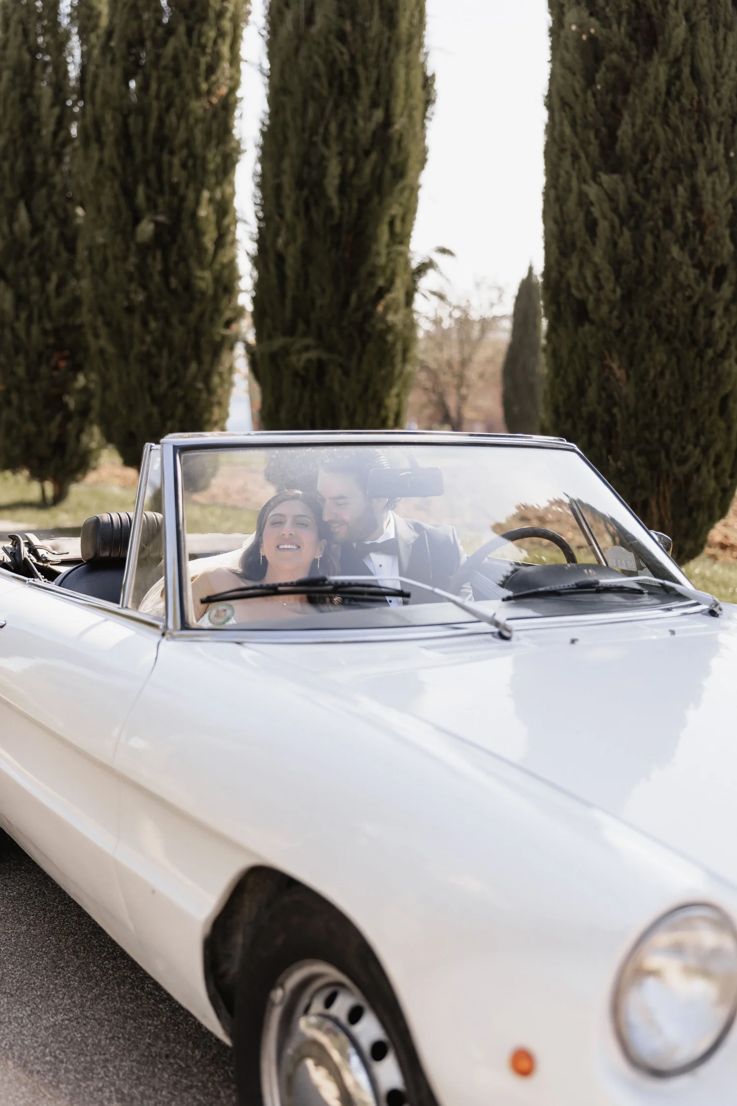 Young couple sitting in a white convertible car, smiling and enjoying a sunny day outdoor with tall trees in the background.