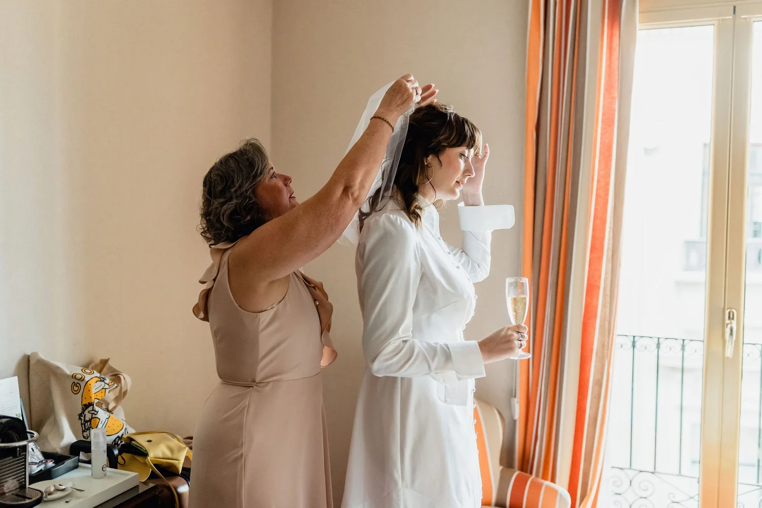 An older woman helping a younger woman put on a veil in a bright room with orange and white curtains, while holding a glass of champagne.