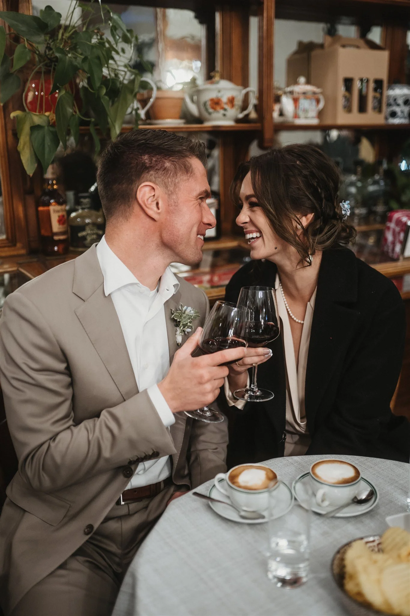 A man and woman smiling and toasting with glasses of red wine at a table containing cappuccinos, a plate of bread, and some water glasses, in a cozy restaurant setting.