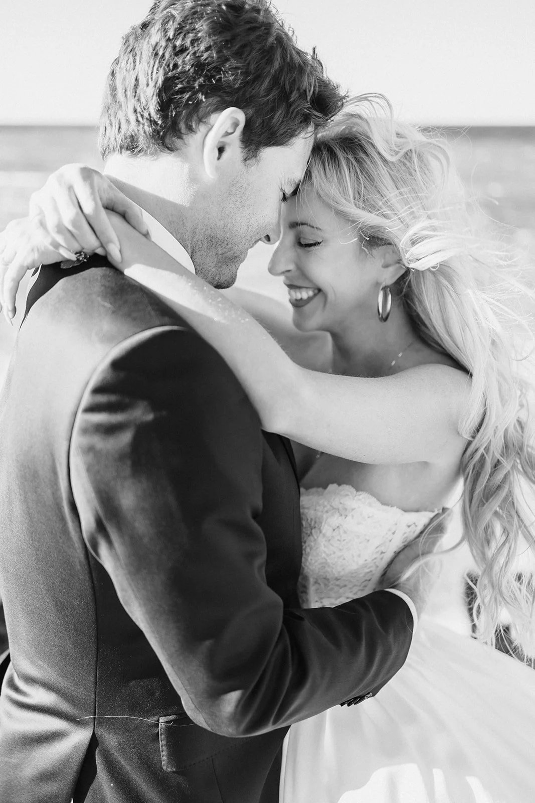 A black and white photo of a happy couple hugging on a beach, smiling with their foreheads touching.