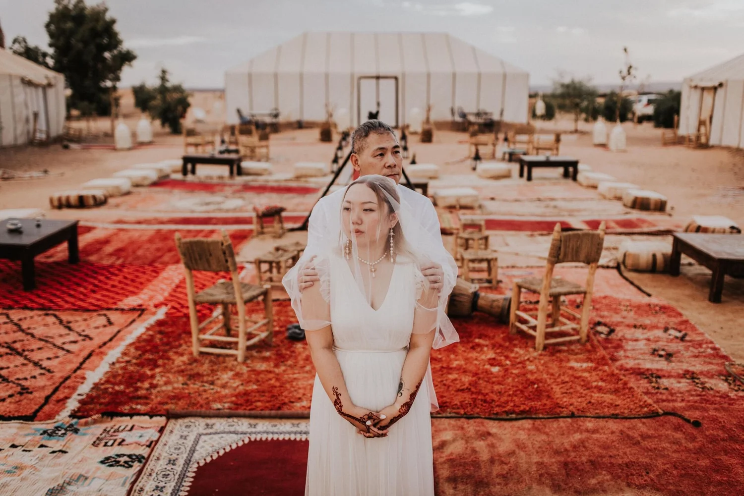 A couple standing outdoors on a desert-like terrain with rugs and furniture, with a tent in the background.