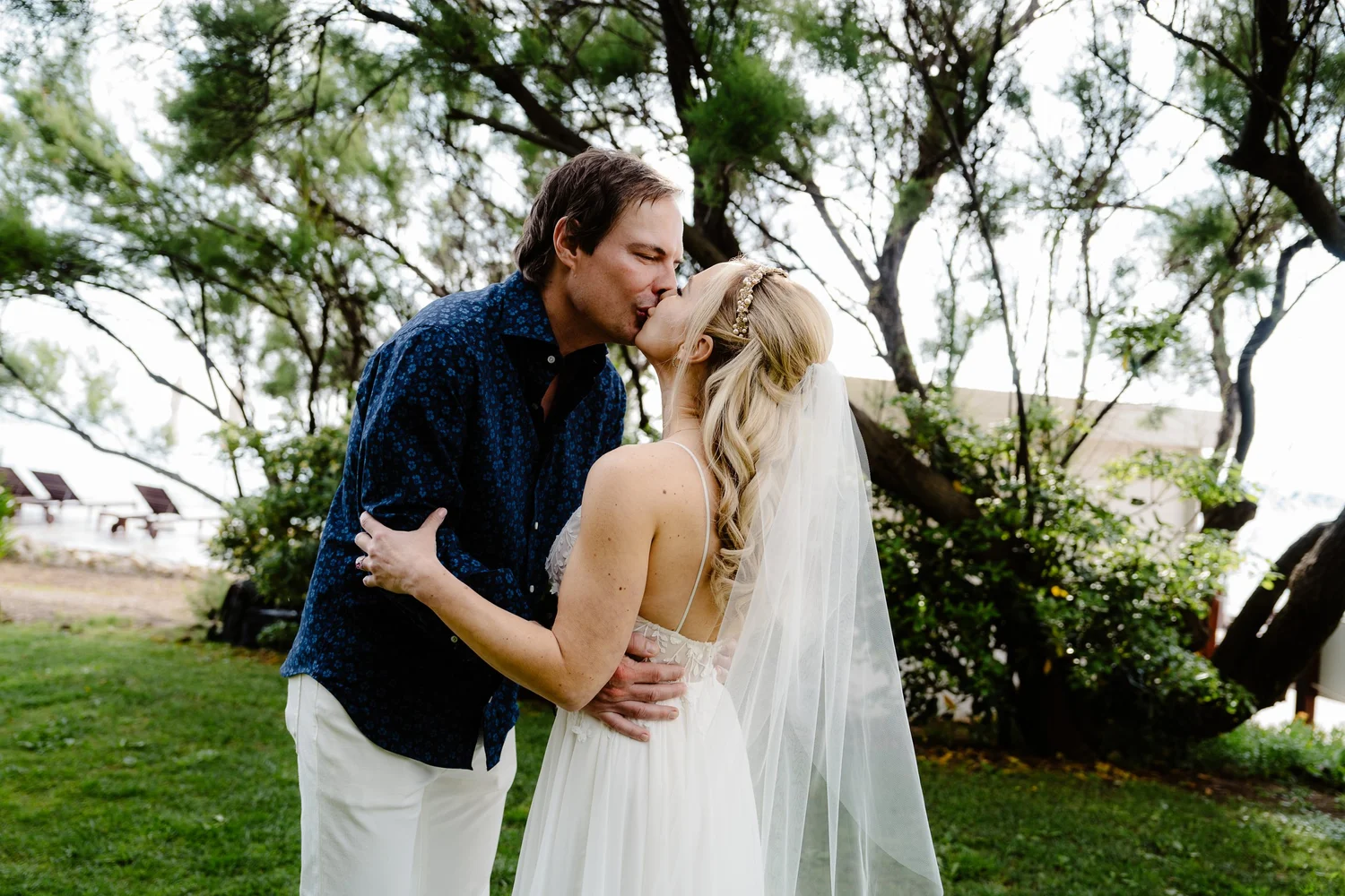 A couple, dressed in wedding attire, sharing a kiss outdoors under a large tree during a wedding ceremony.
