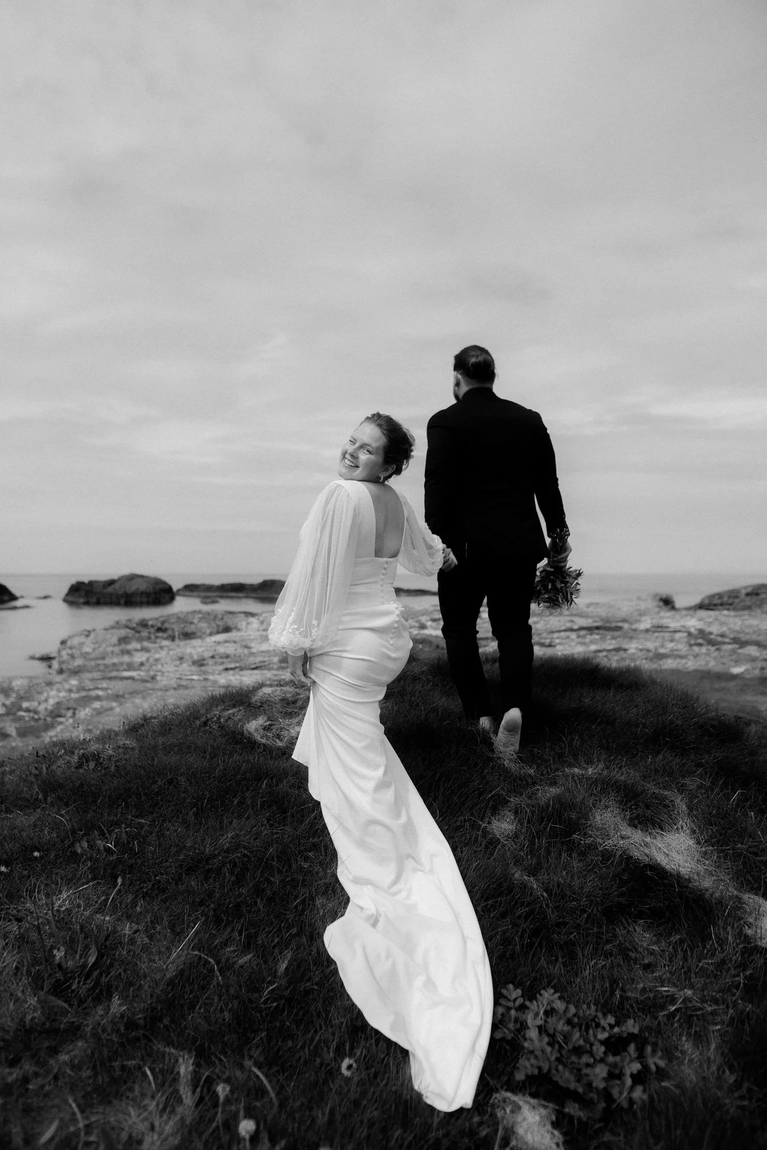 A bride and groom walking hand in hand on a grassy area near rocks and water, with the bride smiling at the camera and the groom holding a bouquet, under a cloudy sky.