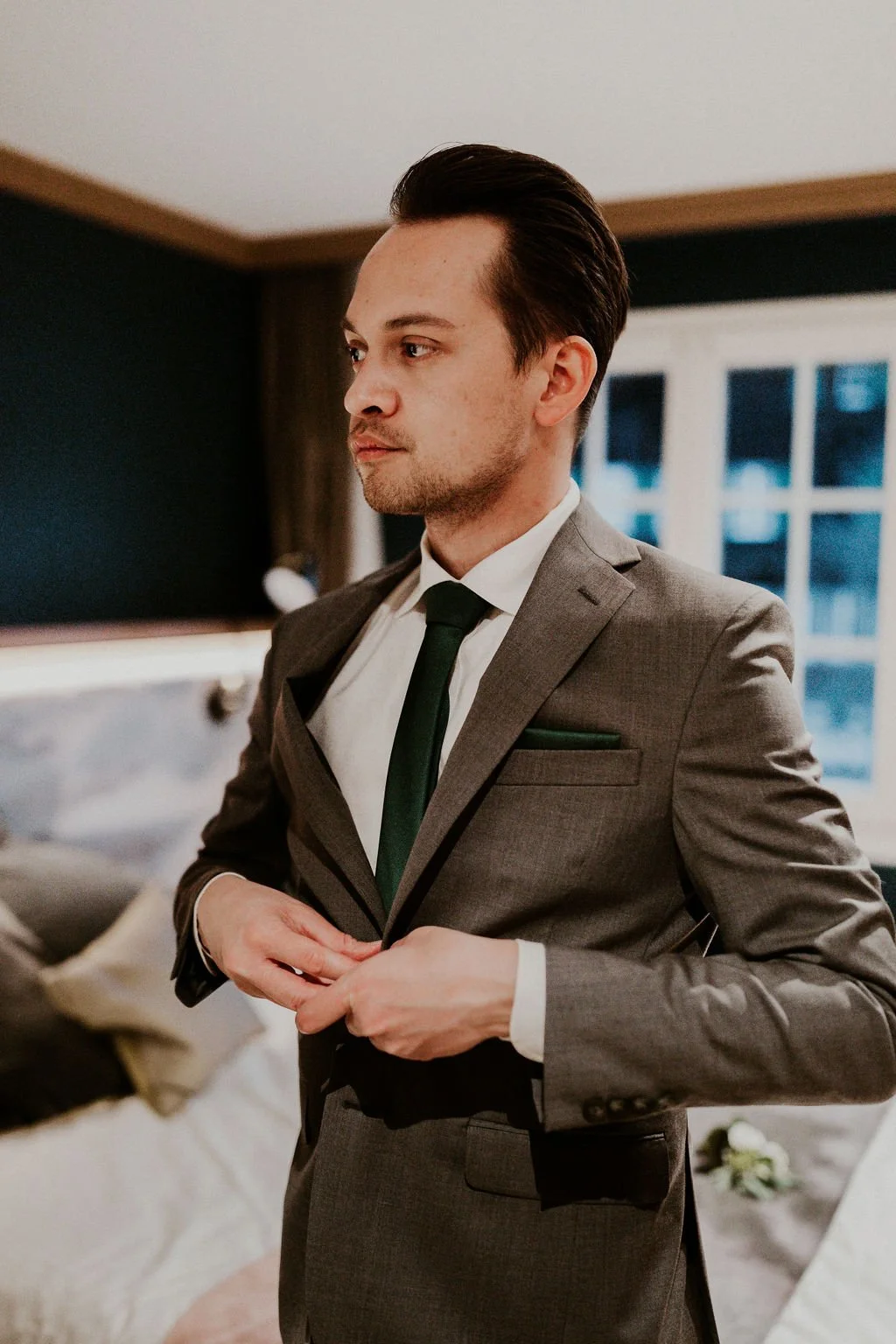 A young man with dark hair combed back, wearing a gray suit, white shirt, and black tie, buttoning his suit jacket indoors with a window in the background.