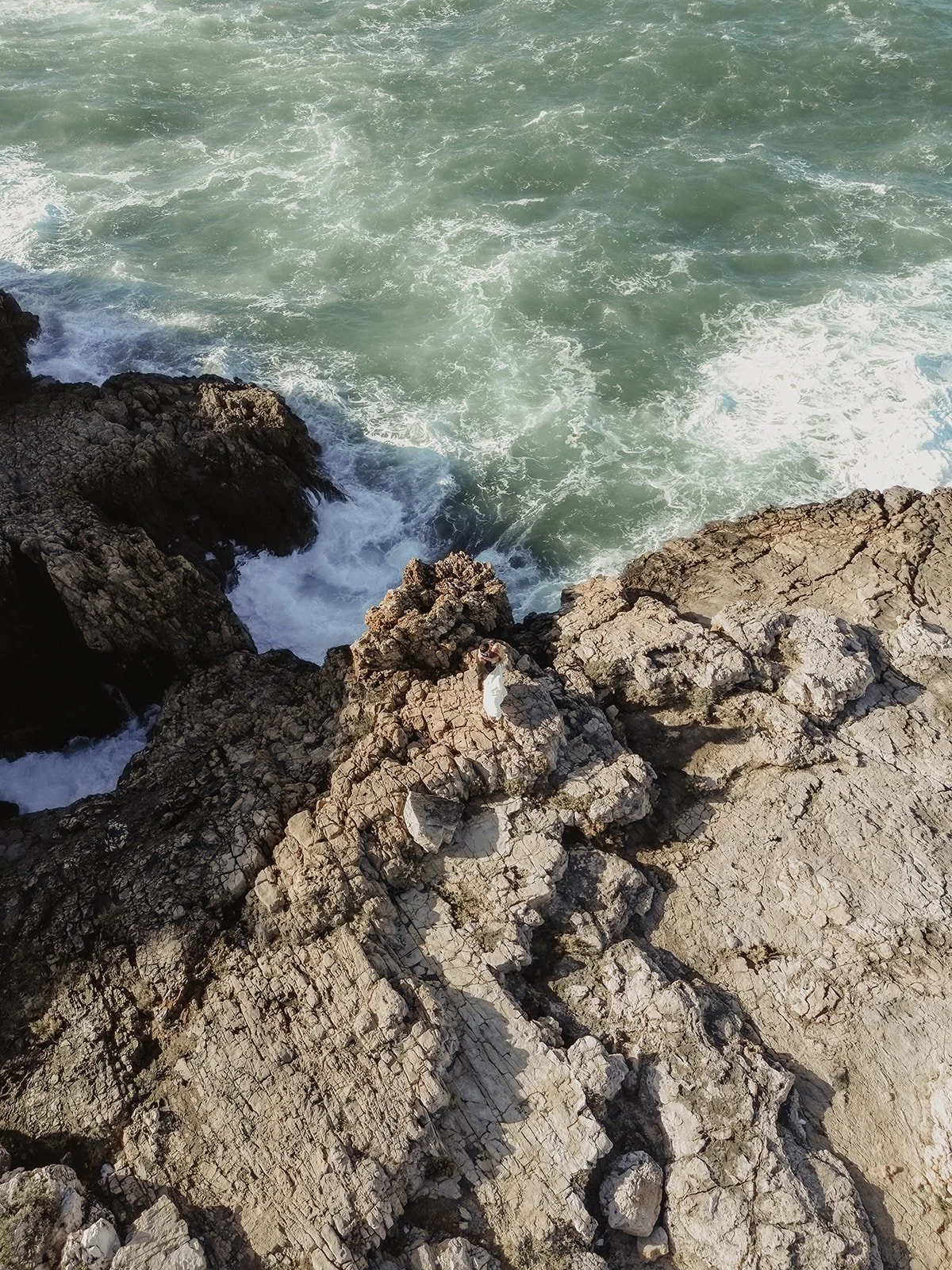 A rocky coastline with waves crashing against the rocks and a person in white clothing standing on one of the rocks.