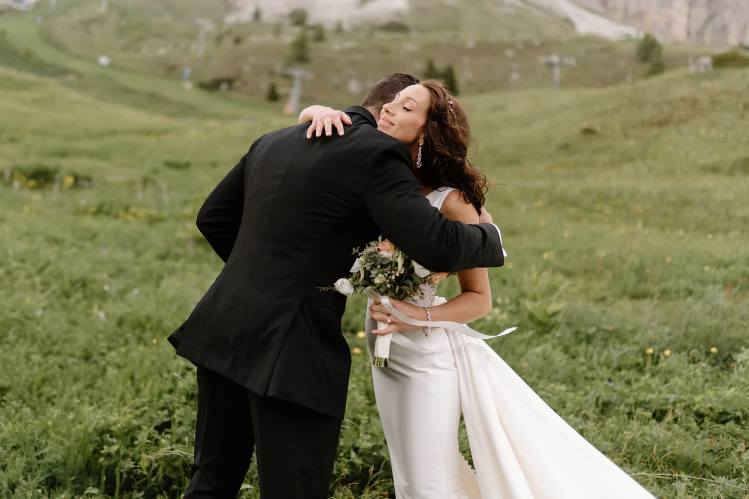 A couple dressed in wedding attire hugging outdoors in a lush green field.
