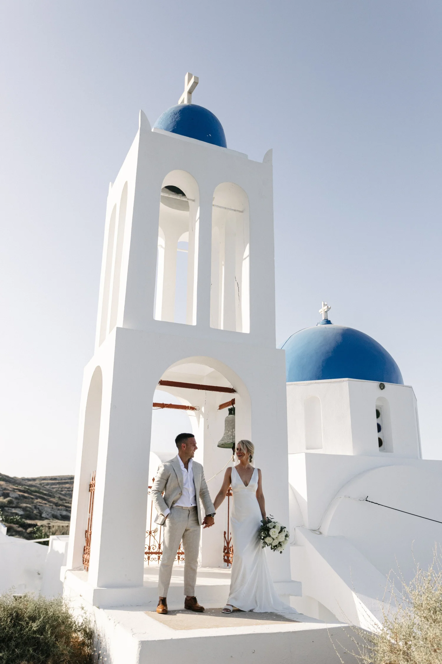 A bride and groom holding hands in front of a white church with blue domes and arches, on a sunny day.