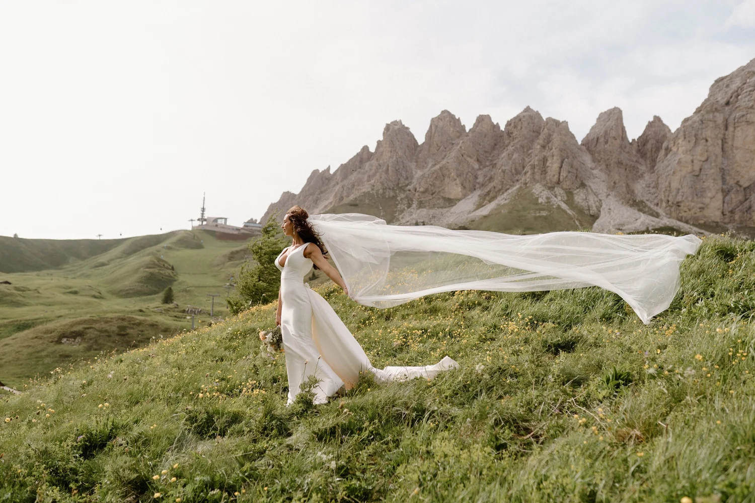 A bride in a white wedding dress with a long flowing veil standing on a grassy hillside with mountains and a communication tower in the background.