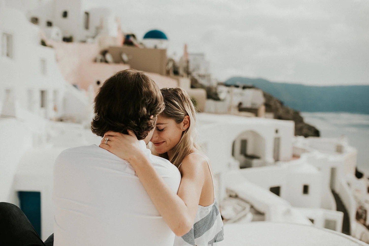 A smiling woman and man sharing an intimate moment outdoors with white buildings and a coastline in the background.