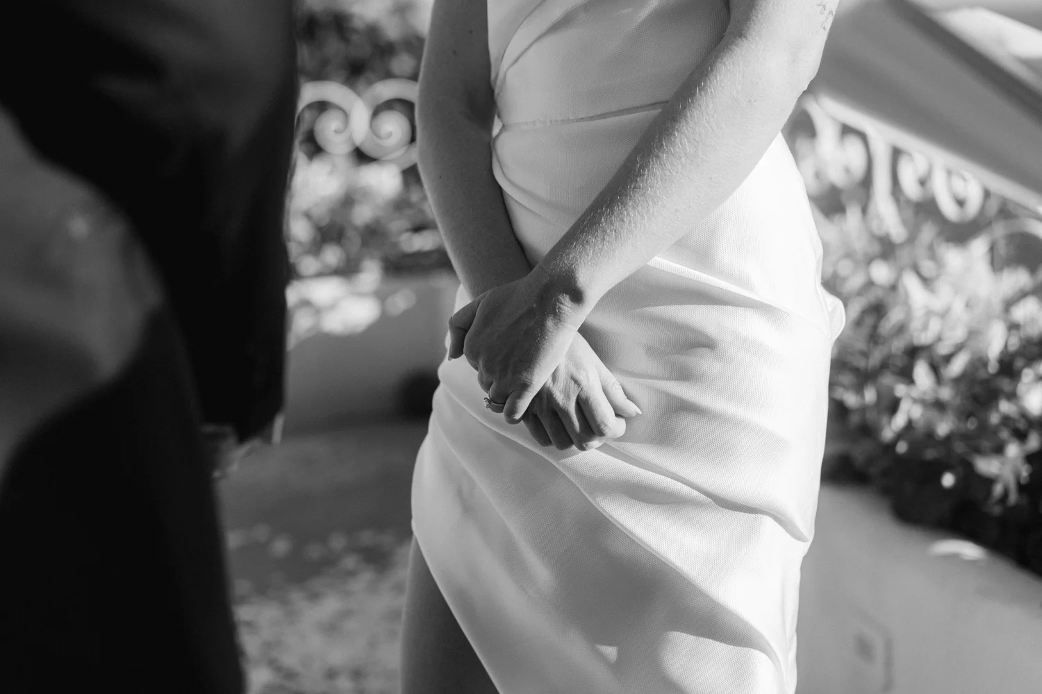 A black and white photo of two people holding hands, with one person wearing a wedding ring, standing outdoors near a decorative white fence.