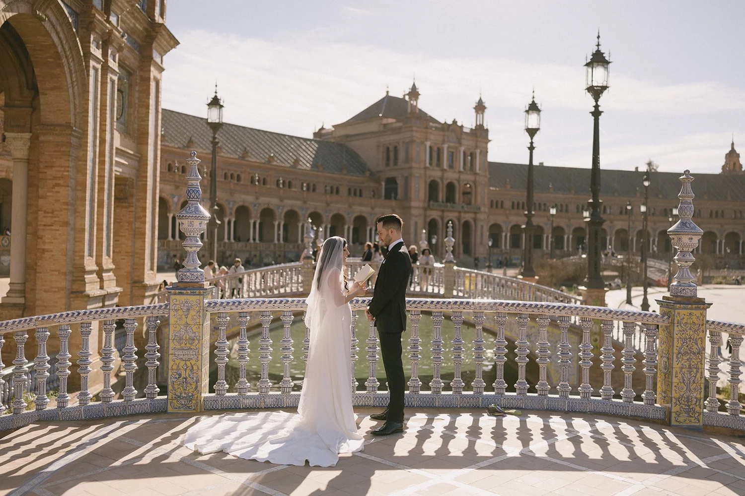 A bride and groom standing on a decorative tile-paved balcony with ornate columns, holding hands and exchanging vows or rings. The bride is wearing a white wedding dress and veil, and the groom is dressed in a black suit. Behind them is a grand, historic building with arches, towers, and decorative lampposts, under a partly cloudy sky.