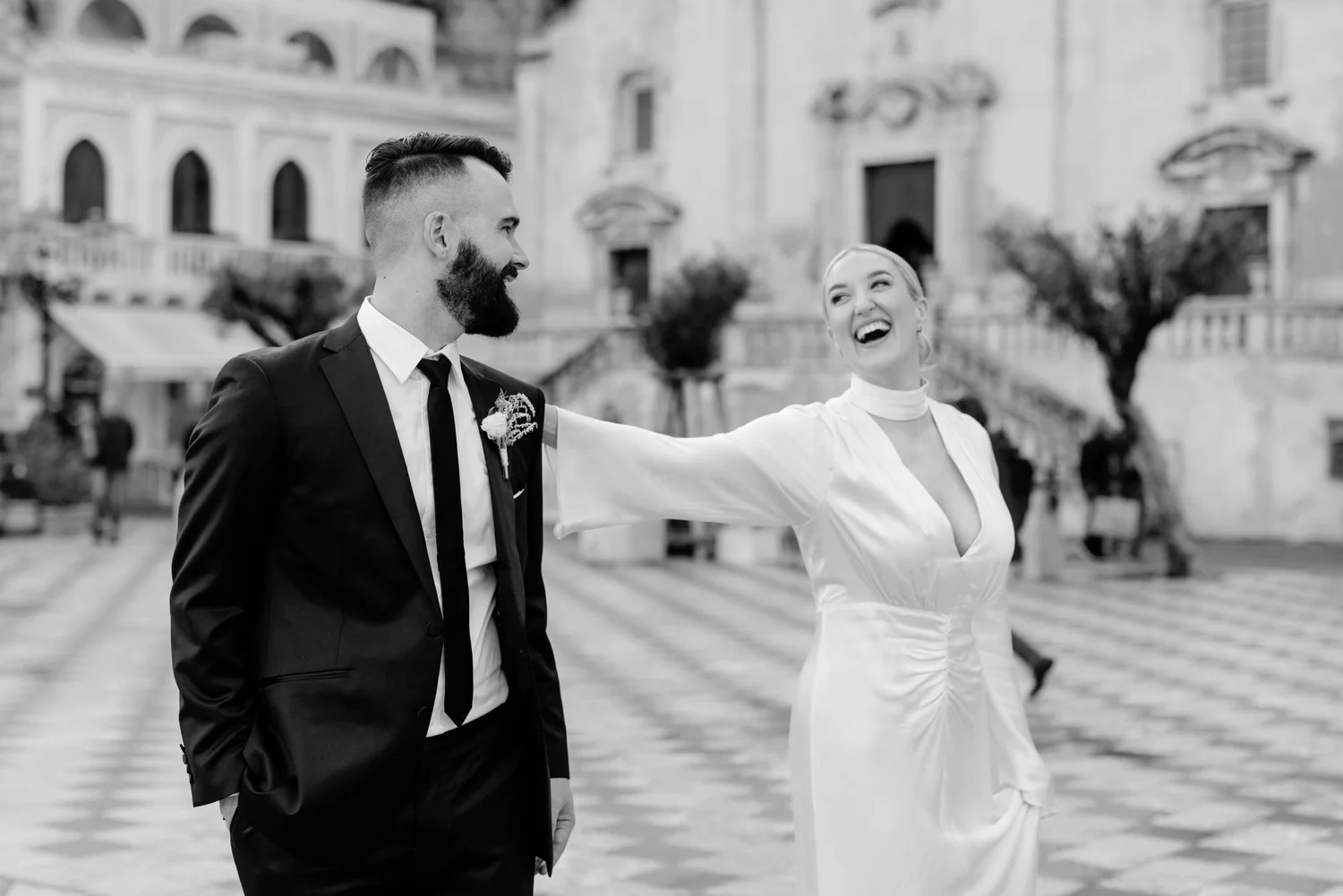A happy couple in formal attire, likely at a wedding, walking outdoors in a European city square with historic buildings in the background. The woman is in a white dress, and the man is in a suit with a boutonniere. They are smiling warmly at each ot