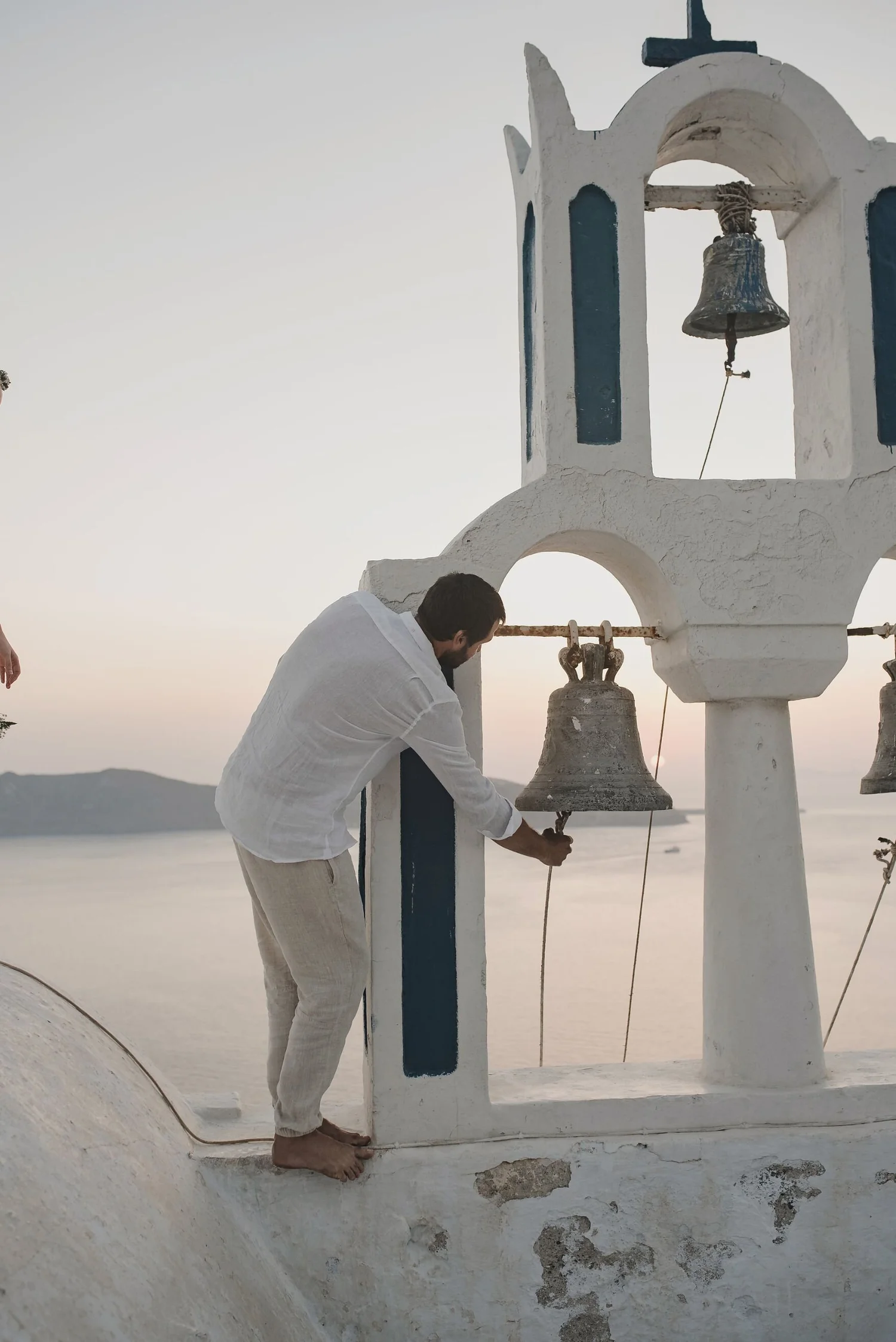 A man wearing a white shirt and beige pants standing barefoot near a white and blue Greek-style bell tower on a hilltop, with a scenic view of the sea and distant landscape in the background during sunset.