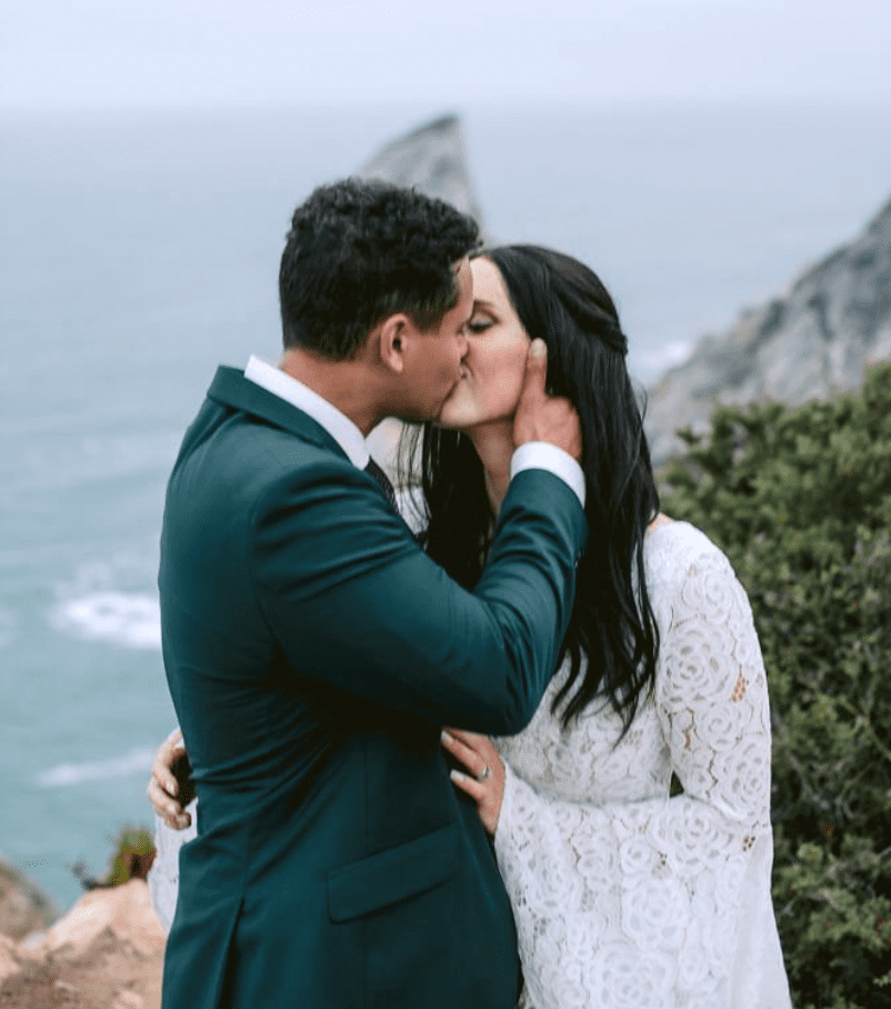 A man and woman kissing outdoors near a rocky coastline and green bushes.