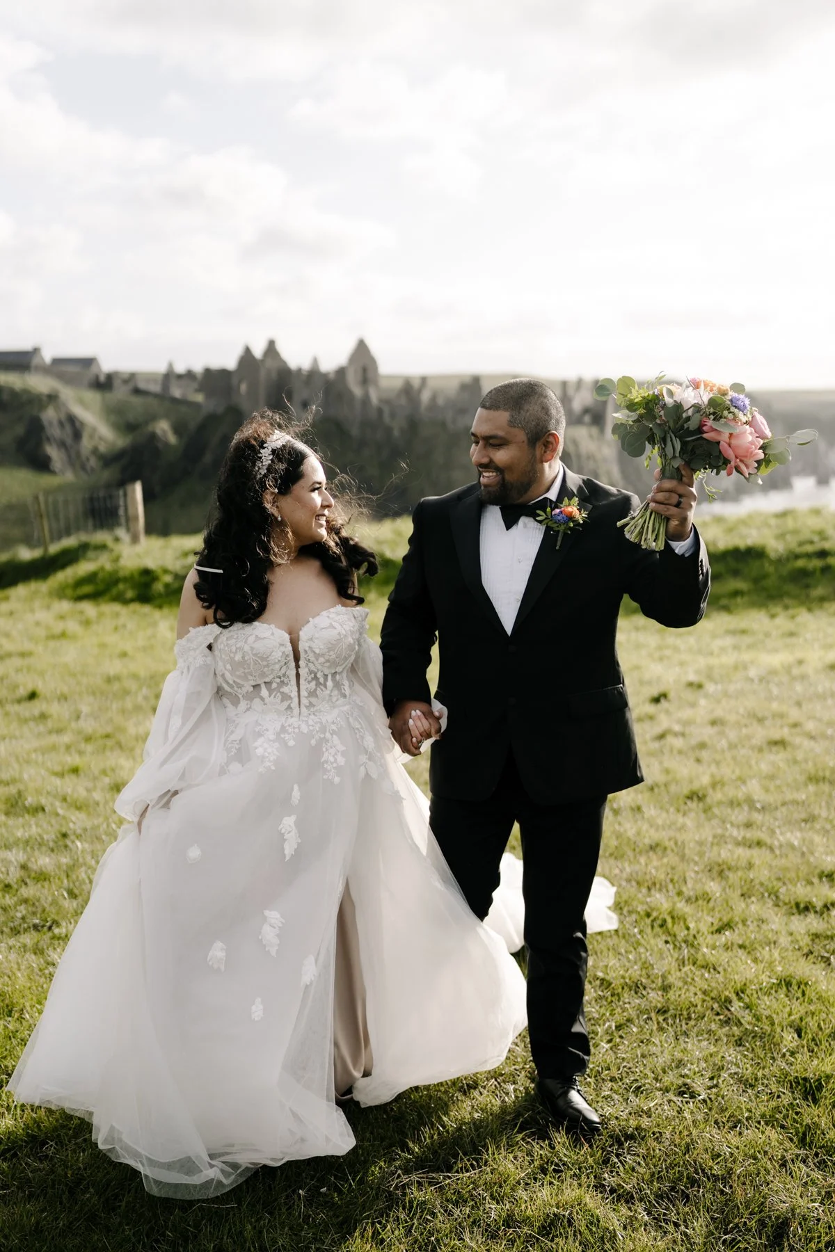 A bride and groom holding hands outdoors, smiling at each other with a castle in the background, the groom holding a bouquet of flowers.