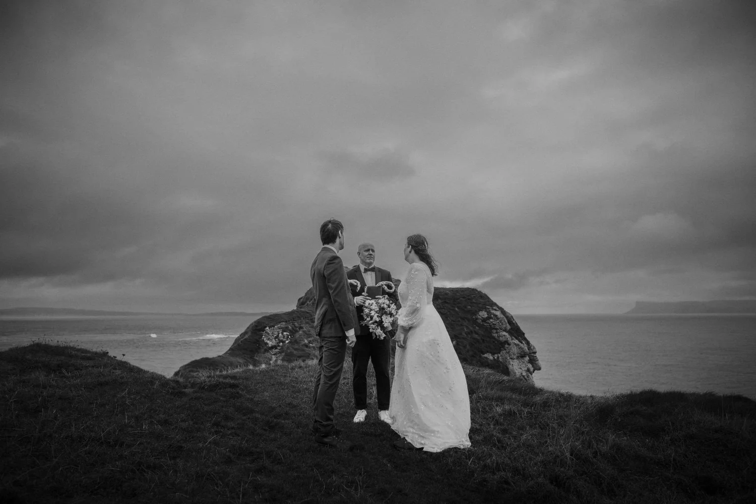 A black and white photograph of a wedding ceremony taking place outdoors on a grassy hill overlooking the ocean, with three individuals in formal attire, including a bride and groom, standing before an officiant.