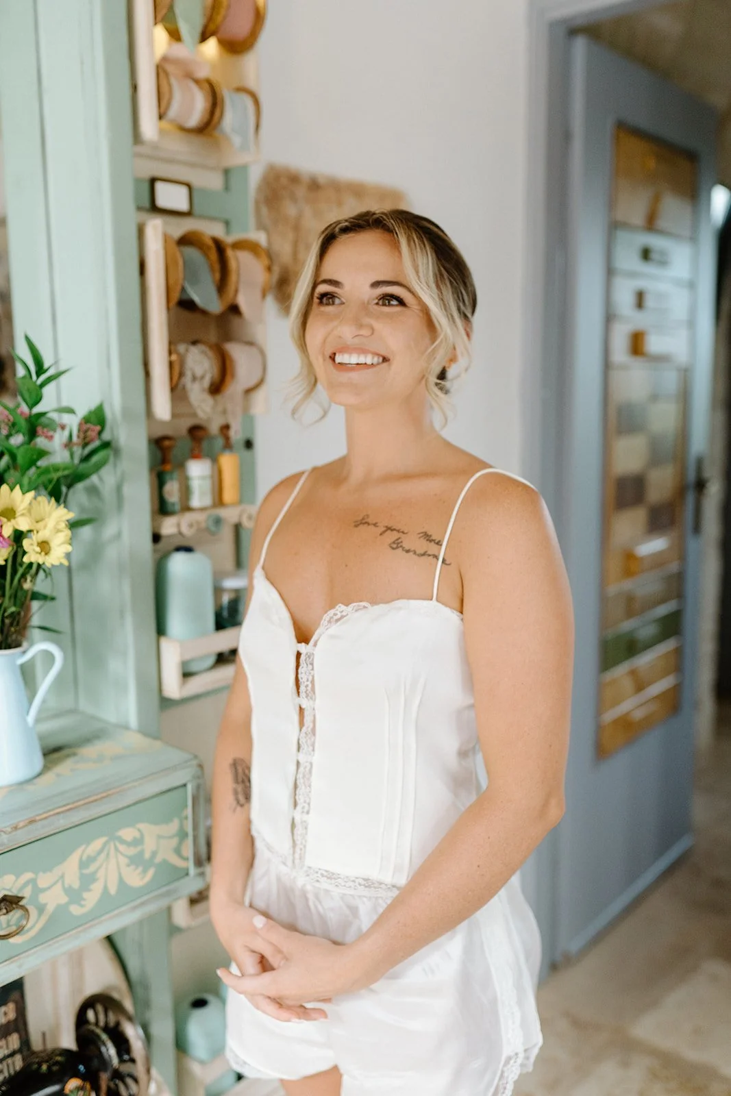 A young woman with short blonde hair and tattoos, wearing a white spaghetti strap nightgown, standing indoors near a green cabinet with flowers and kitchen items, smiling.